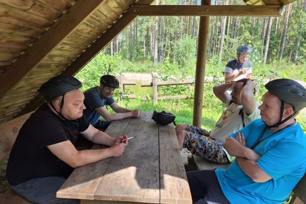 Mieszkańcy siedza pod wiatą leśną w kaskach rowerowych.