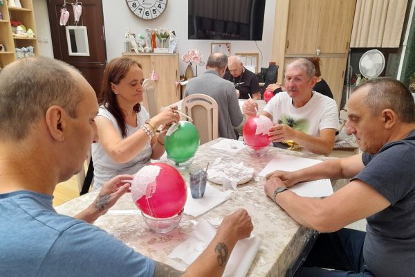 Mieszkańcy przyklejaja papierki na balony siedząc przy stolikach w sali terapii zajęciowej.