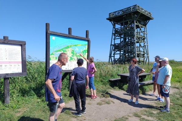 Mieszkańcy przy tablicy informacyjnej a na drugim planie wieża widokowa.