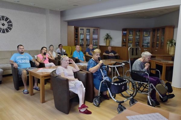 Kilkoro mieszkańców siedzi w fotelach w kawiarence i śpiewa.