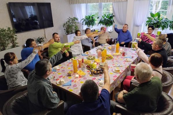 Uczestnicy imprezy urodzinowej wznoszą toast siedząc w fotelach na około dużego stołu z poczęstunkiem.