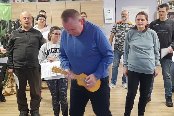 Mieszkaniec wśród kilkoro innych uczestników próby wokalnej improwizuje grę na mandolinie.