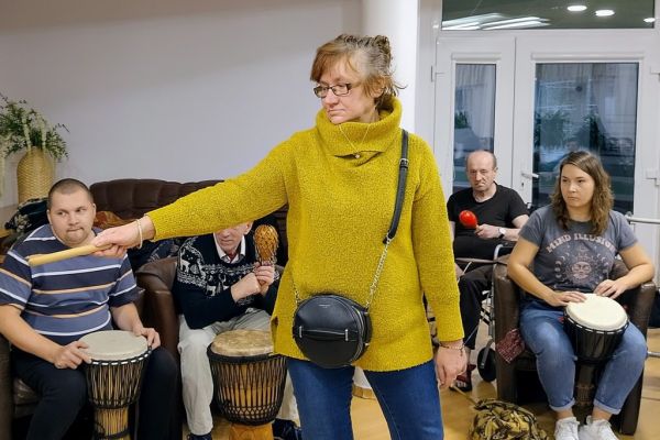 Mieszkanka stojąc wskazuje ręką kolejnego gracza wśród siedzących w koło.