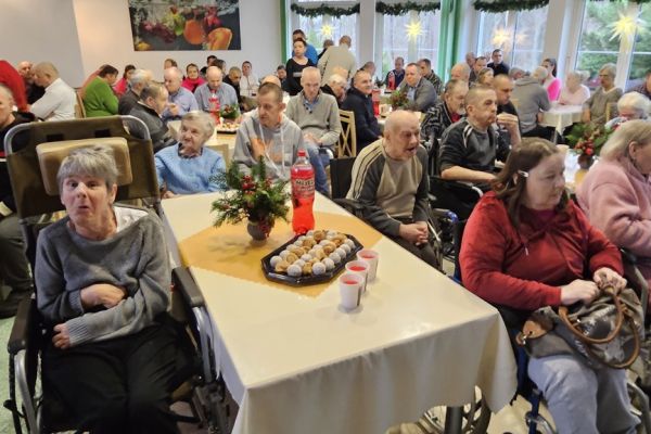 Dużo mieszkańców siedzi przy stołach z poczęstunkiem na stołówce.