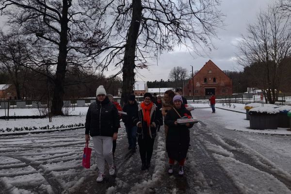 Grupa mieszkańców z panią Dyrektor idzie po dworzu w zomowym krajobrazie.