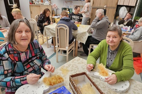 Mieszkańcy w sali terapii siedzą i wykonują różne prace manualne przy stolikach.