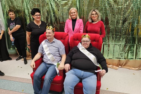 Dwoje uczestników balu w koronach i szarfach siedza na fotelach a za nimi stoją trzy panie