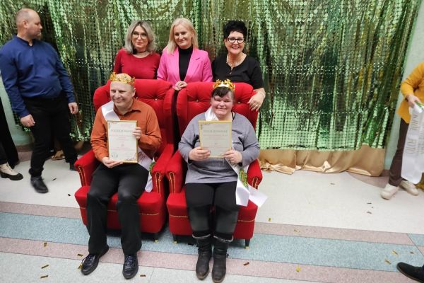 Dwoje uczestników balu w koronach i szarfach siedza na fotelach a za nimi stoją trzy panie