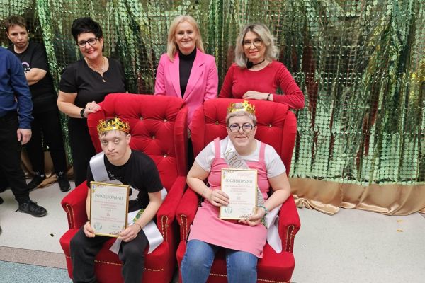 Dwoje uczestników balu w koronach i szarfach siedza na fotelach a za nimi stoją trzy panie