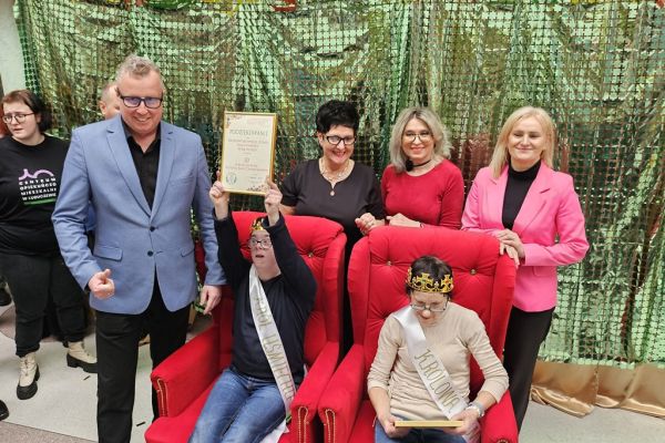 Dwoje uczestników balu w koronach i szarfach siedza na fotelach a za nimi stoją trzy panie