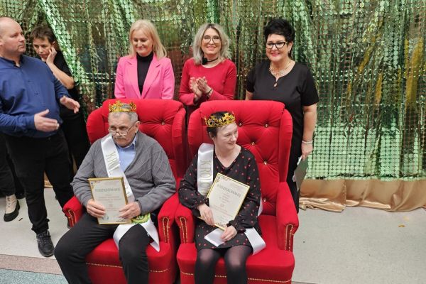 Dwoje uczestników balu w koronach i szarfach siedza na fotelach a za nimi stoją trzy panie