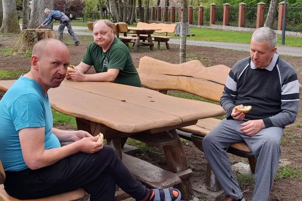 Mieszkańcy siedza na ławeczce i spożywają kiełbaskę.