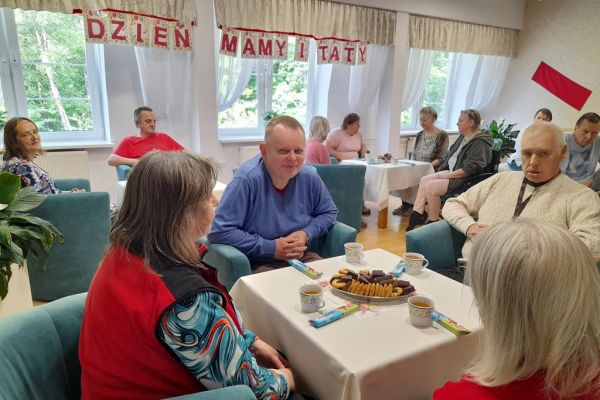 Mieszkańcy siedzą przy stolikach z poczęstunkiem w kawiarence.