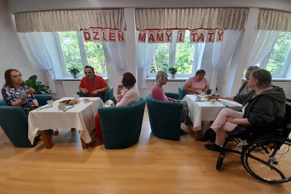 Mieszkańcy siedzą przy stolikach z poczęstunkiem w kawiarence.