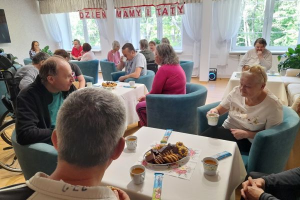 Mieszkańcy siedzą przy stolikach z poczęstunkiem w kawiarence.