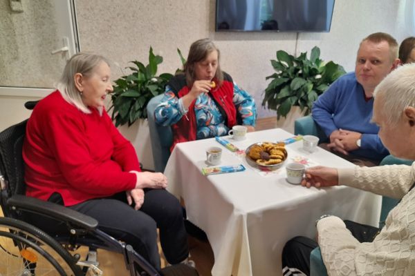 Mieszkańcy siedzą przy stolikach z poczęstunkiem w kawiarence.