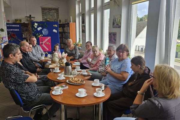 Mieszkańcy i inni uczestnicy spotkania siedza w bibliotece przy stolikach i słuchaja tekstu czytanego.