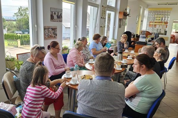 Mieszkańcy i inni uczestnicy spotkania siedza w bibliotece przy stolikach i słuchaja tekstu czytanego.