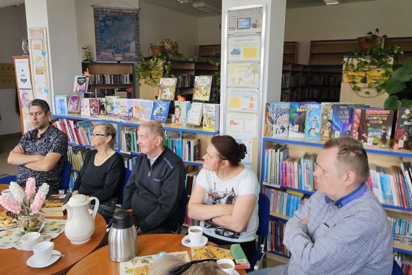 Mieszkańcy i inni uczestnicy spotkania siedza w bibliotece przy stolikach i słuchaja tekstu czytanego.