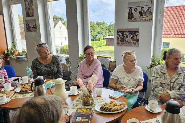 Mieszkańcy i inni uczestnicy spotkania siedza w bibliotece przy stolikach i słuchaja tekstu czytanego.