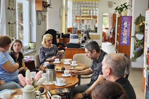 Mieszkańcy i inni uczestnicy spotkania siedza w bibliotece przy stolikach i słuchaja tekstu czytanego.