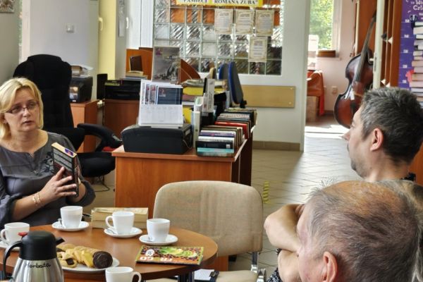 Mieszkańcy i inni uczestnicy spotkania siedza w bibliotece przy stolikach i słuchaja tekstu czytanego.