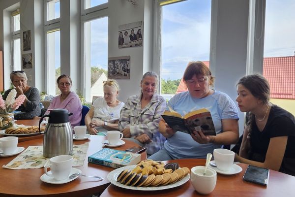 Mieszkańcy i inni uczestnicy spotkania siedza w bibliotece przy stolikach i słuchaja tekstu czytanego.